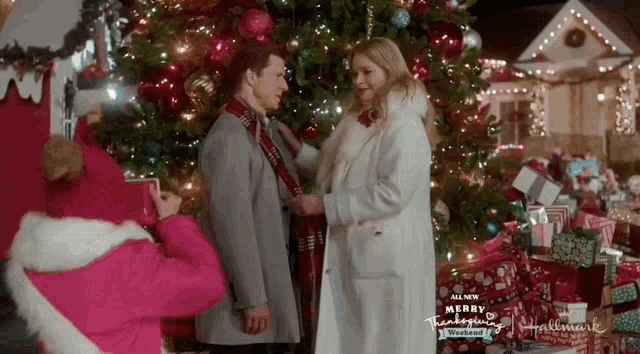 a man and woman standing in front of a christmas tree with a sign that says merry christmas on it
