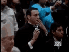 a man in a tuxedo is sitting in a crowd of people with the letter g on the bottom right corner