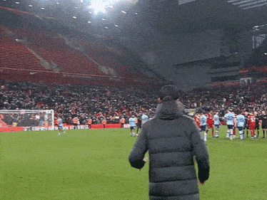 a man in a black jacket stands on a soccer field in front of a large crowd