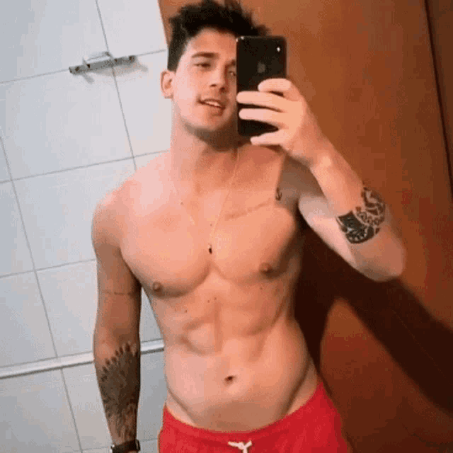 a shirtless man in red shorts is taking a selfie with his phone