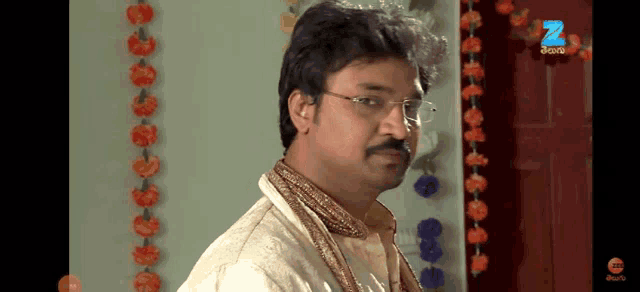 a man with glasses and a mustache is standing in front of a door with flowers hanging from it and the letter z on it