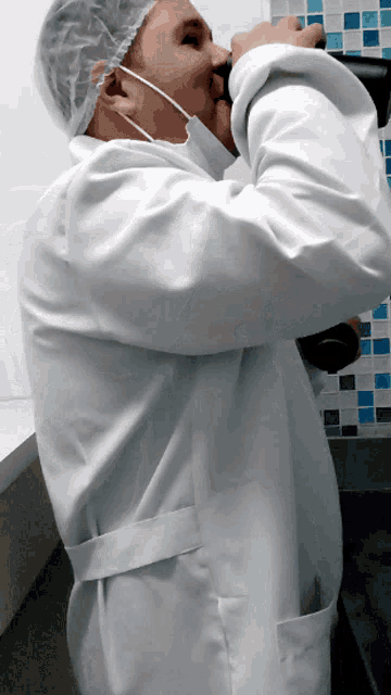 a man wearing a white lab coat and a plastic cap is drinking from a bottle
