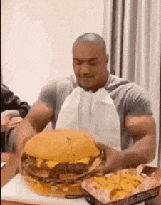 a man is eating a very large hamburger and french fries at a table .