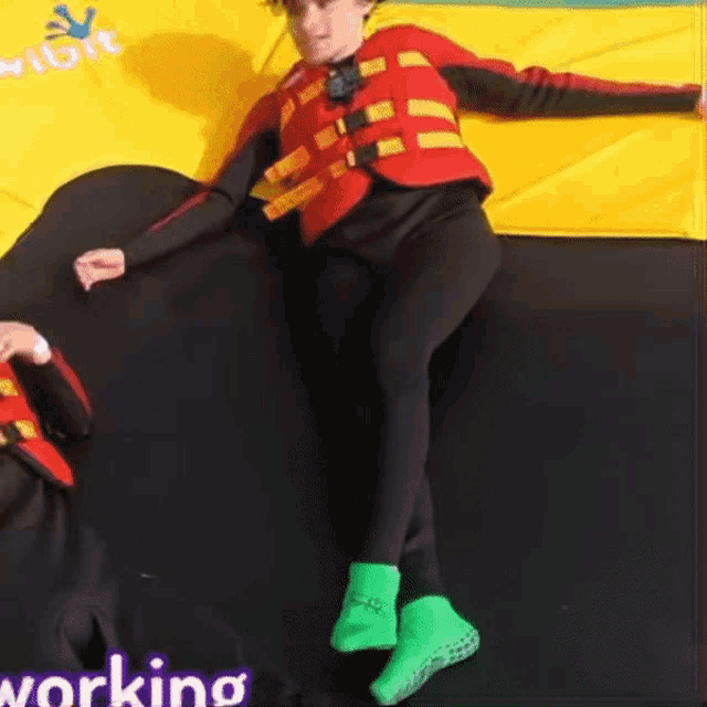 a person wearing a life jacket and green socks is laying on a trampoline with the word working in the lower right corner
