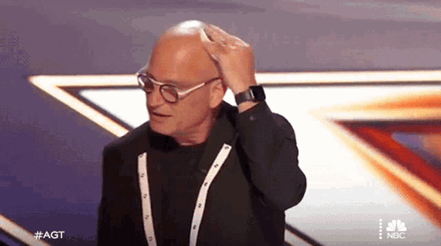 a bald man wearing glasses and a watch is standing on a stage with the nbc logo in the background