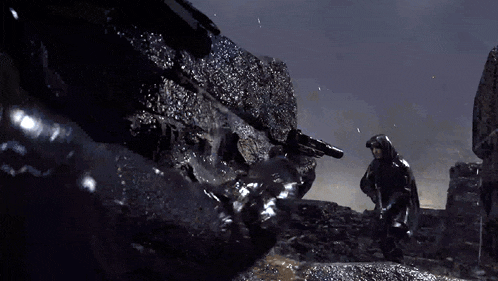 a man in a hooded jacket is holding a gun in front of a large rock .
