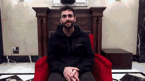 a man in a black hoodie sits in a red chair with his hands folded