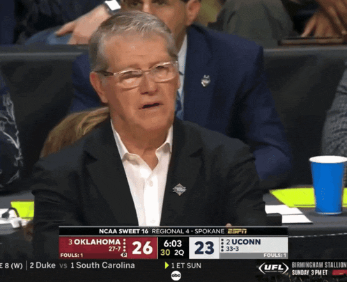 a man in a suit sits in the stands watching a ncaa basketball game between oklahoma and uconn