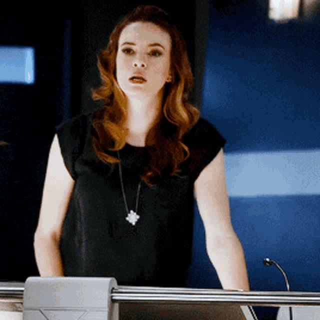 a woman in a black top and necklace stands in front of a microphone