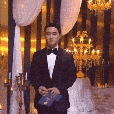 a man in a tuxedo is standing in front of a chandelier holding a blue book