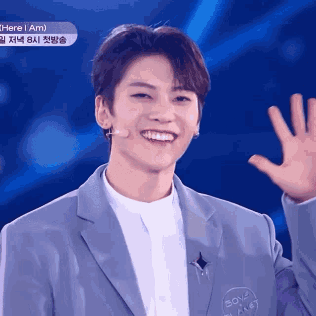 a young man in a suit is smiling and waving his hand in front of a sign that says here i am