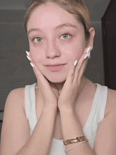 a woman with white nails and a gold bracelet on her wrist touches her face