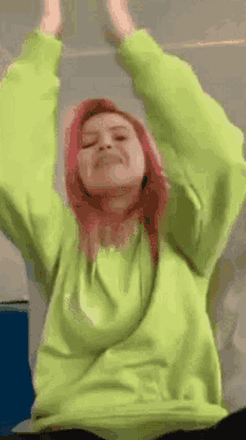 a woman with pink hair is wearing a neon green sweatshirt and raising her arms in the air .