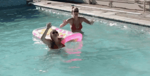 two women are playing in a swimming pool and one is floating on a pink raft .