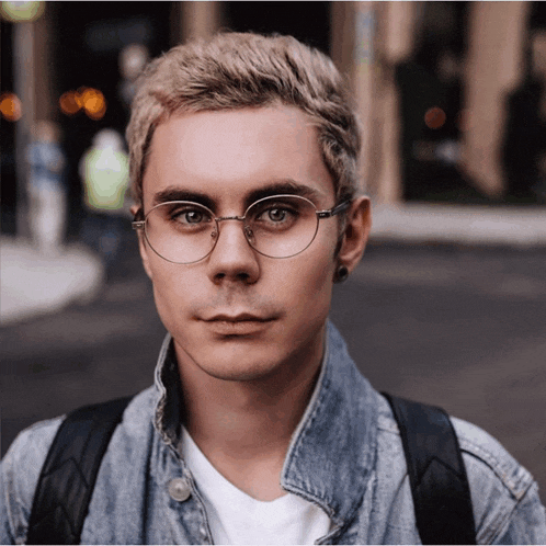 a young man wearing glasses and a denim jacket is looking at the camera