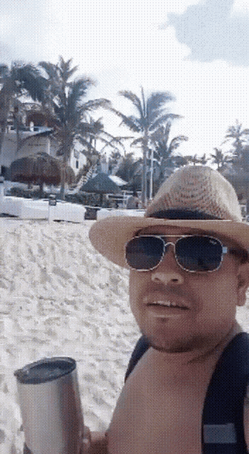 a man wearing a straw hat and sunglasses is holding a tumbler on the beach