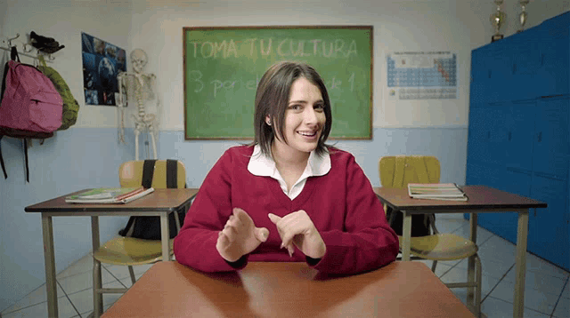 a woman in a red sweater sits at a desk in front of a blackboard that says toma tu cultura