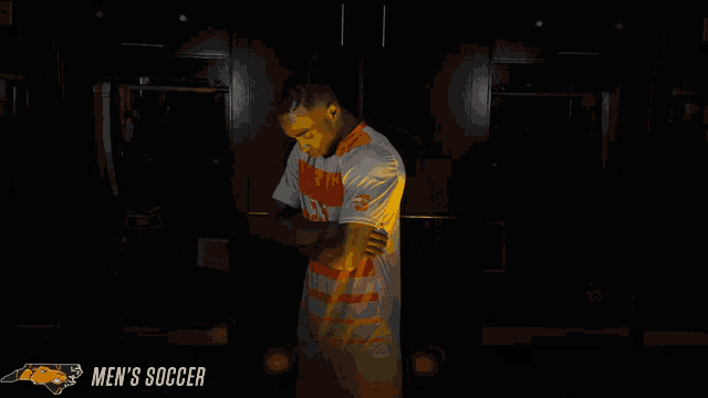 a man in an orange and white striped shirt is standing in a dark room with the words men 's soccer below him