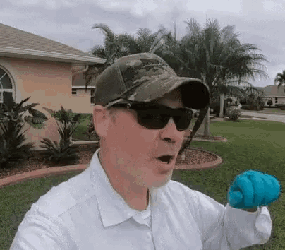a man wearing a hat and sunglasses is standing in front of a house holding a blue glove .