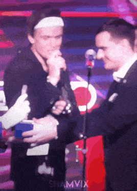 a man in a headband is talking into a microphone while a man in a suit shakes hands with him