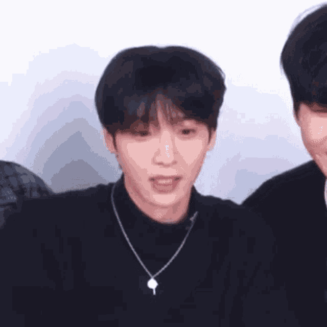 a young man wearing a black sweater and a necklace with a key on it is sitting next to another young man .
