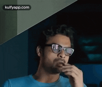 a man wearing glasses and a blue shirt is smoking a cigarette in a dark room .