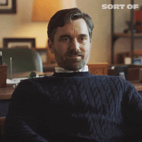 a man in a blue sweater is sitting in front of a desk with the word sort of written on the bottom