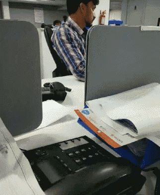 a man in a plaid shirt sits at a desk behind a cubicle divider