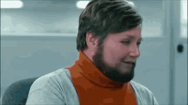 a man with a beard wearing an orange turtleneck is sitting in an office .