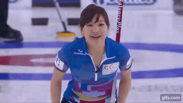 a woman in a blue shirt is smiling while standing on a curling rink .