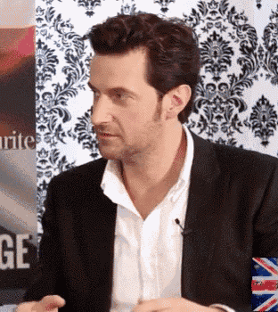 a man in a suit and white shirt is sitting in front of a wall with a pattern .