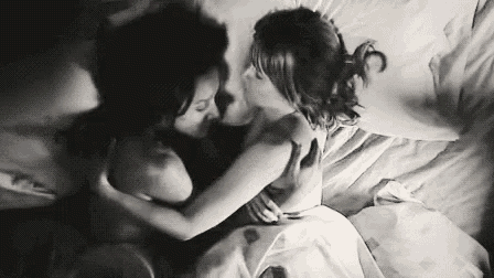 a black and white photo of two women kissing while laying on a bed .