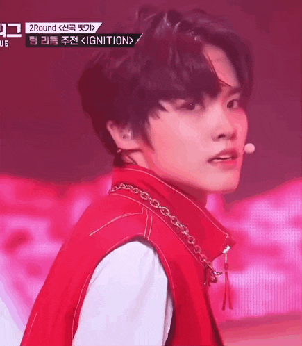 a young man wearing a red vest and a chain around his neck is standing in front of a microphone on a stage .
