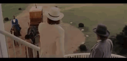a man in a white hat is standing on a balcony looking at a man riding a horse .