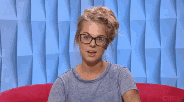 a woman wearing glasses is sitting on a pink couch with a cbs logo in the background