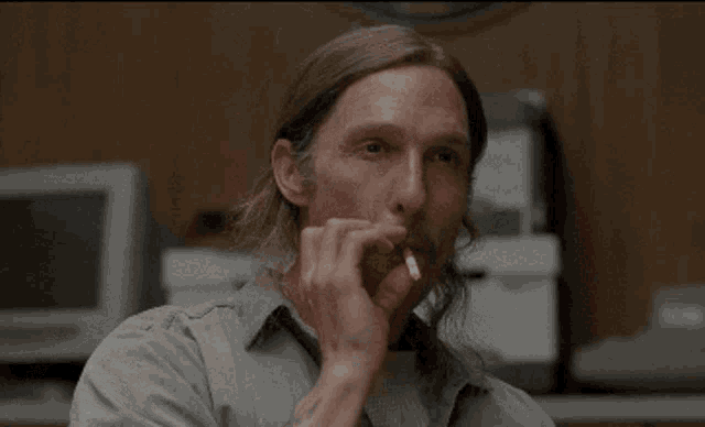 a man with long hair and a mustache is smoking a cigarette and saying shee-it