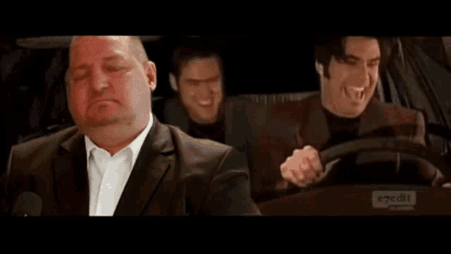 a man in a suit is sleeping in the back seat of a car while two other men are driving .