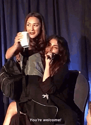two women are sitting in front of a blue curtain and one of them is saying you 're welcome ..