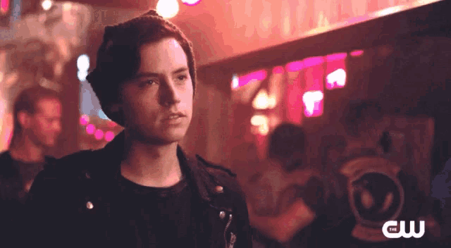 a man wearing a beanie and a leather jacket is standing in front of a cw logo