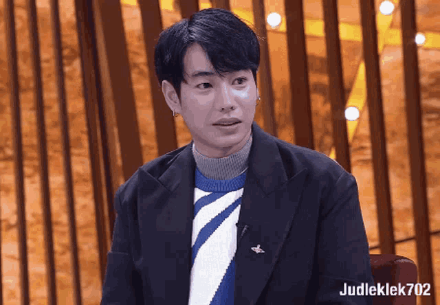 a man in a suit and sweater is sitting in front of a wooden wall with the name judleklek702 on the bottom right