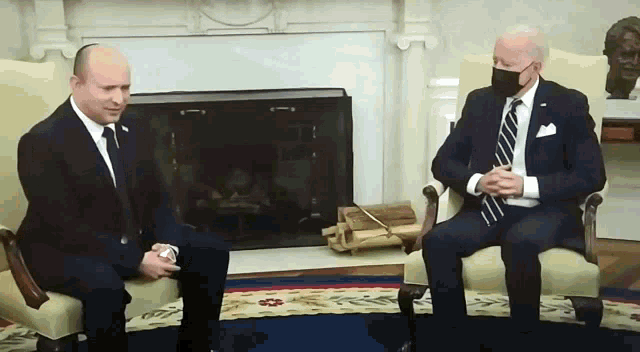 two men wearing suits and ties are sitting in chairs in front of a fireplace