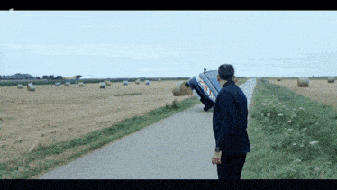 a man stands on the side of a road looking at a car that is on its side in a field