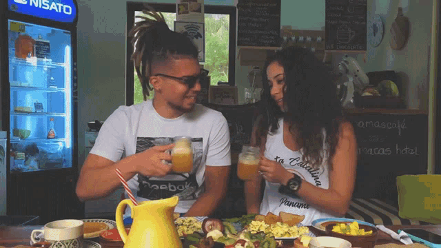 a man and a woman sit at a table with a nisato fridge in the background