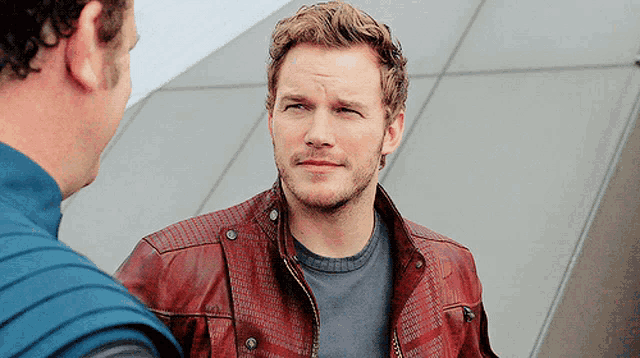 a man with a beard wearing a red leather jacket talks to another man