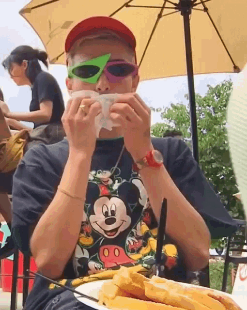 a man wearing mickey mouse sunglasses is eating a hot dog