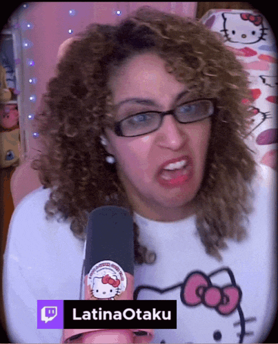 a woman with curly hair wearing glasses and a hello kitty shirt holds a microphone and says latinaotaku