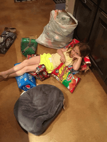 a little girl is laying on the floor surrounded by bags of chips and a bag of pepsi