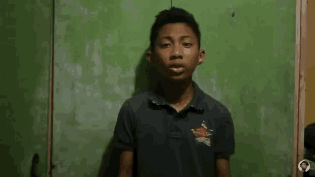 a young boy is standing in front of a green wall and making a gesture with his hands that says seen .