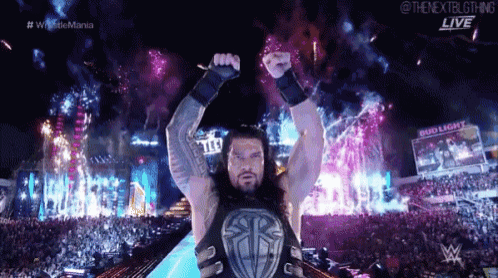 roman reigns is standing in front of a crowd holding his arms in the air .