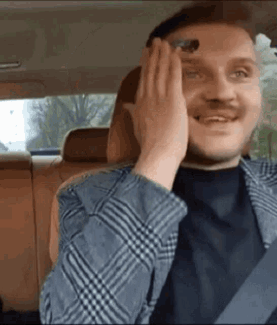 a man in a plaid jacket is sitting in the back seat of a car covering his face with his hands .